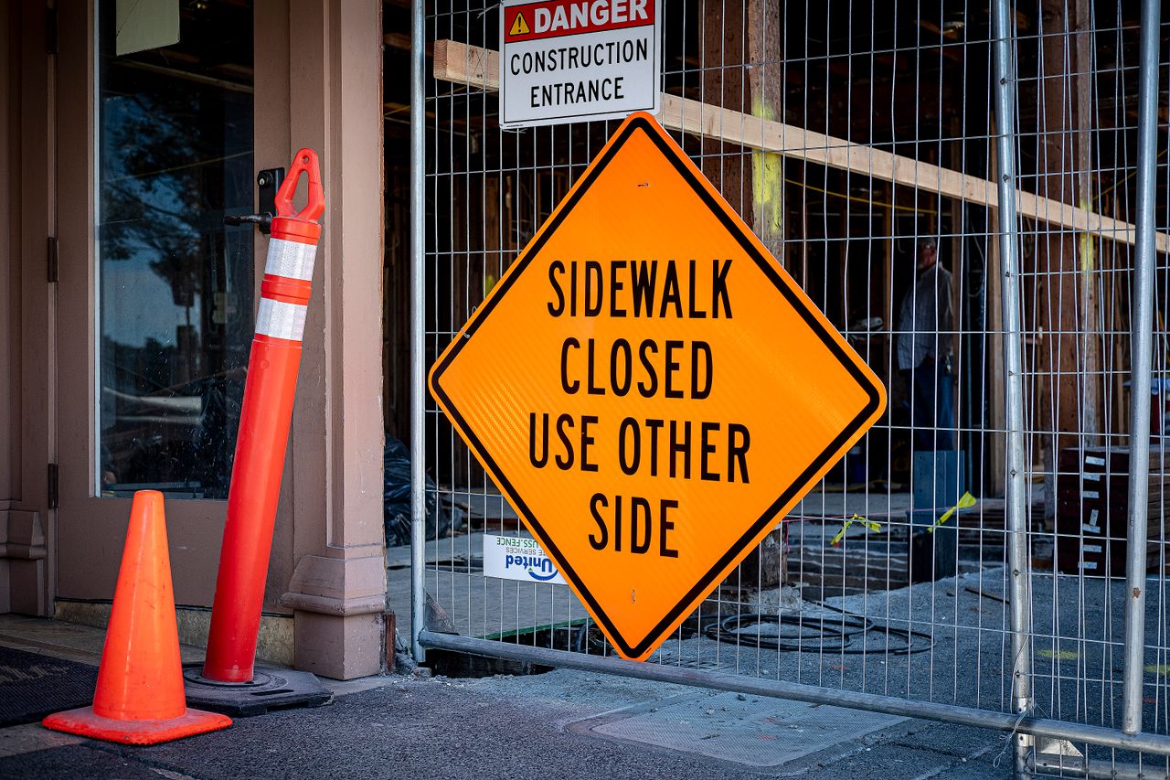 about-us Bright orange warning sign indicating sidewalk closure at construction entrance.